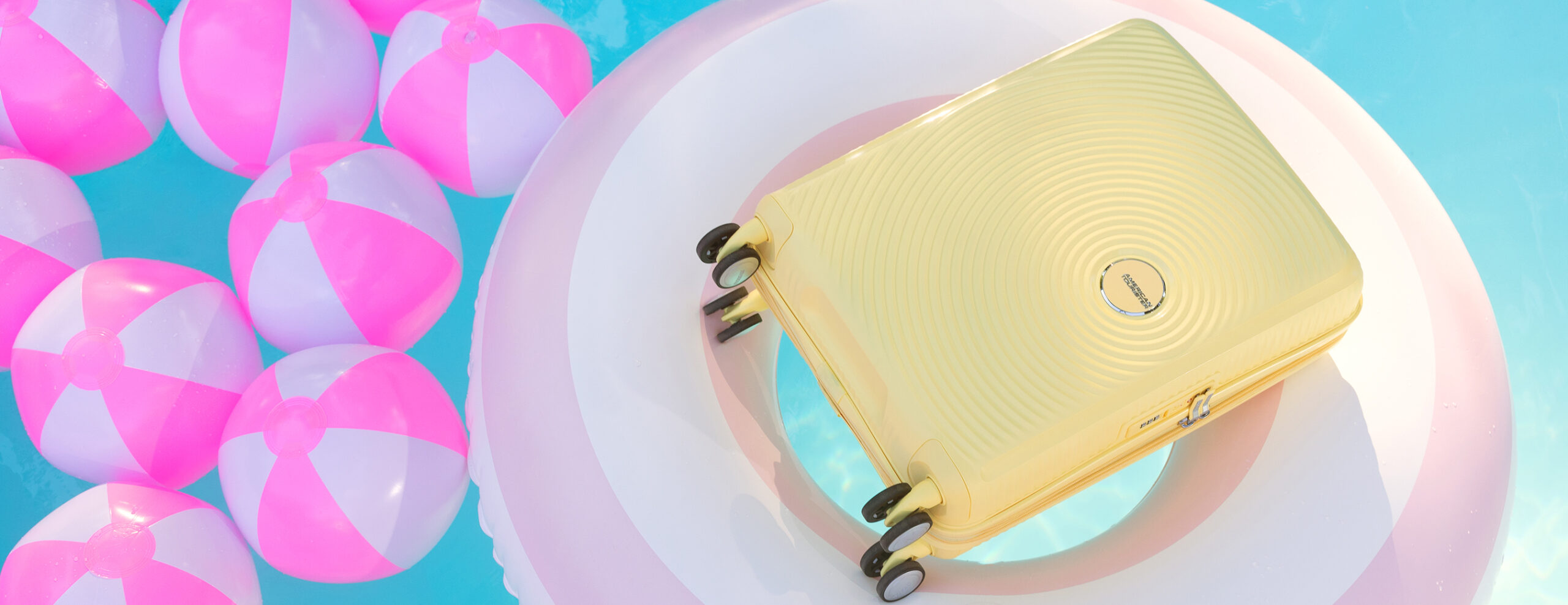 Sommergave: pastellgul American Tourister Soundbox-koffert på flytering i basseng med rosa badeballer