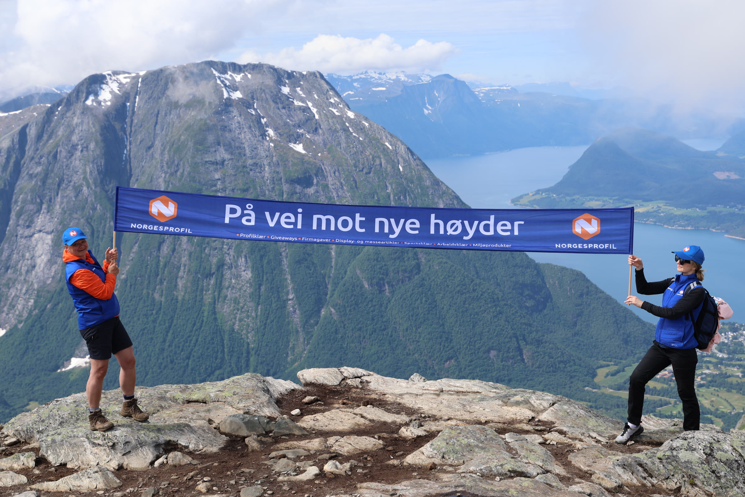 To ansatte i Norgesprofil holder banneret «På vei mot nye høyder» på en fjelltopp med utsikt over fjorden