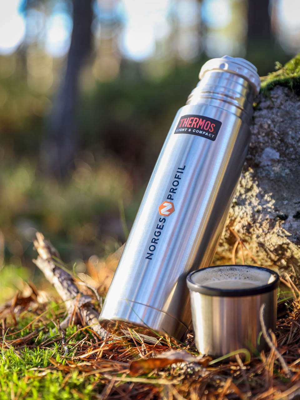 Termos fra Thermos i børstet stål med Norgesprofil-logo, stående på mose i skogen ved siden av en kopp kaffe